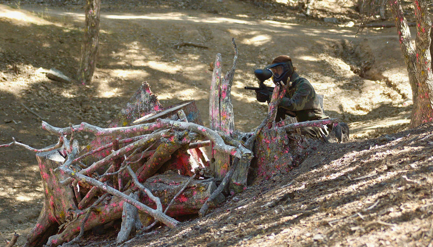 A person crouched behind a pile of branches in the middle of a paintball game.