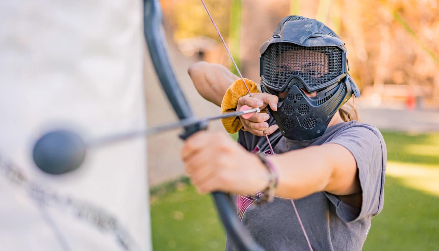 A woman in archery tag gear, aiming a bow and arrow.